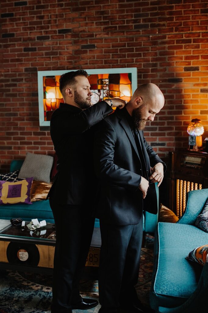 A groom looks down while adjusting his suit coat as one of his groomsmen behind him adjusts the suit coat collar