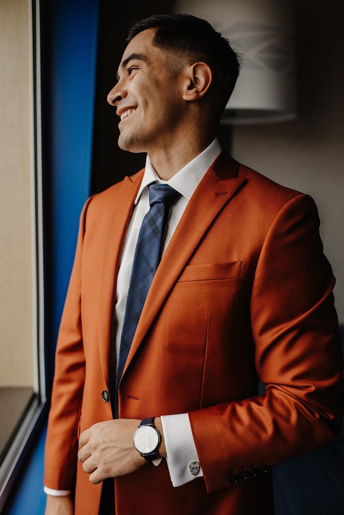 A groom in an orange suit smiles to the left while looking out a window