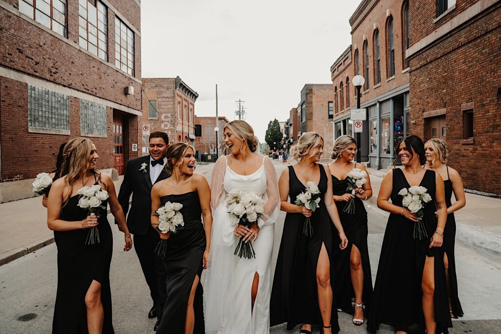 A bride in the middle of her wedding party of 8 walk down a street of Chicago together while laughing with one another, on either side of them are brick buildings