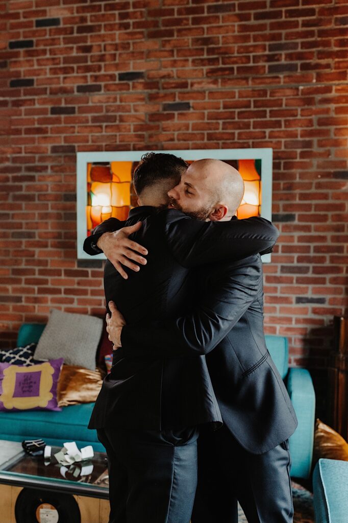 A groom hugs one of his groomsmen before his wedding day as the two get ready in a brick room