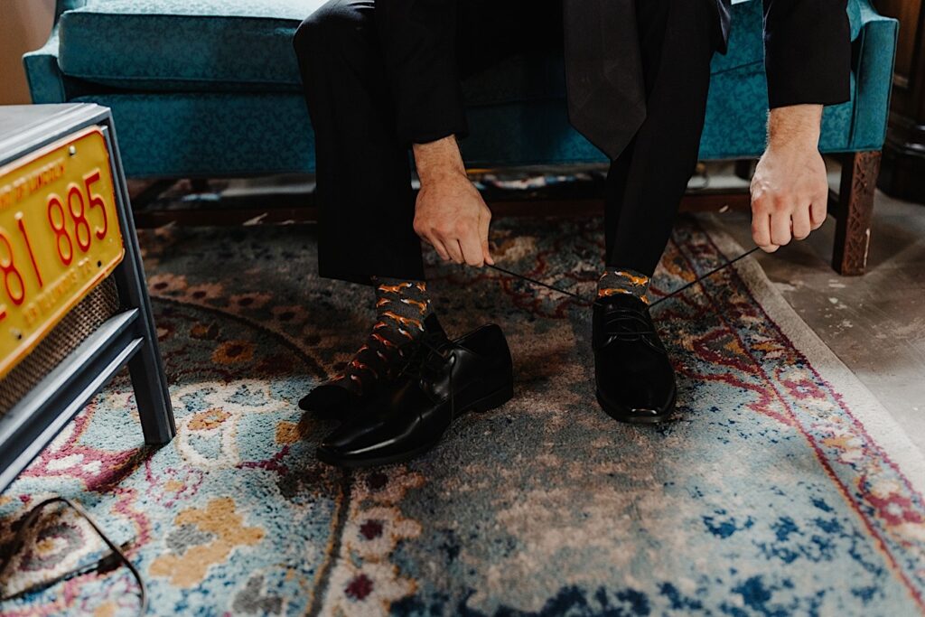 Knee down photo of a groom sitting on a blue couch tying his shoelaces while wearing socks with foxes on them