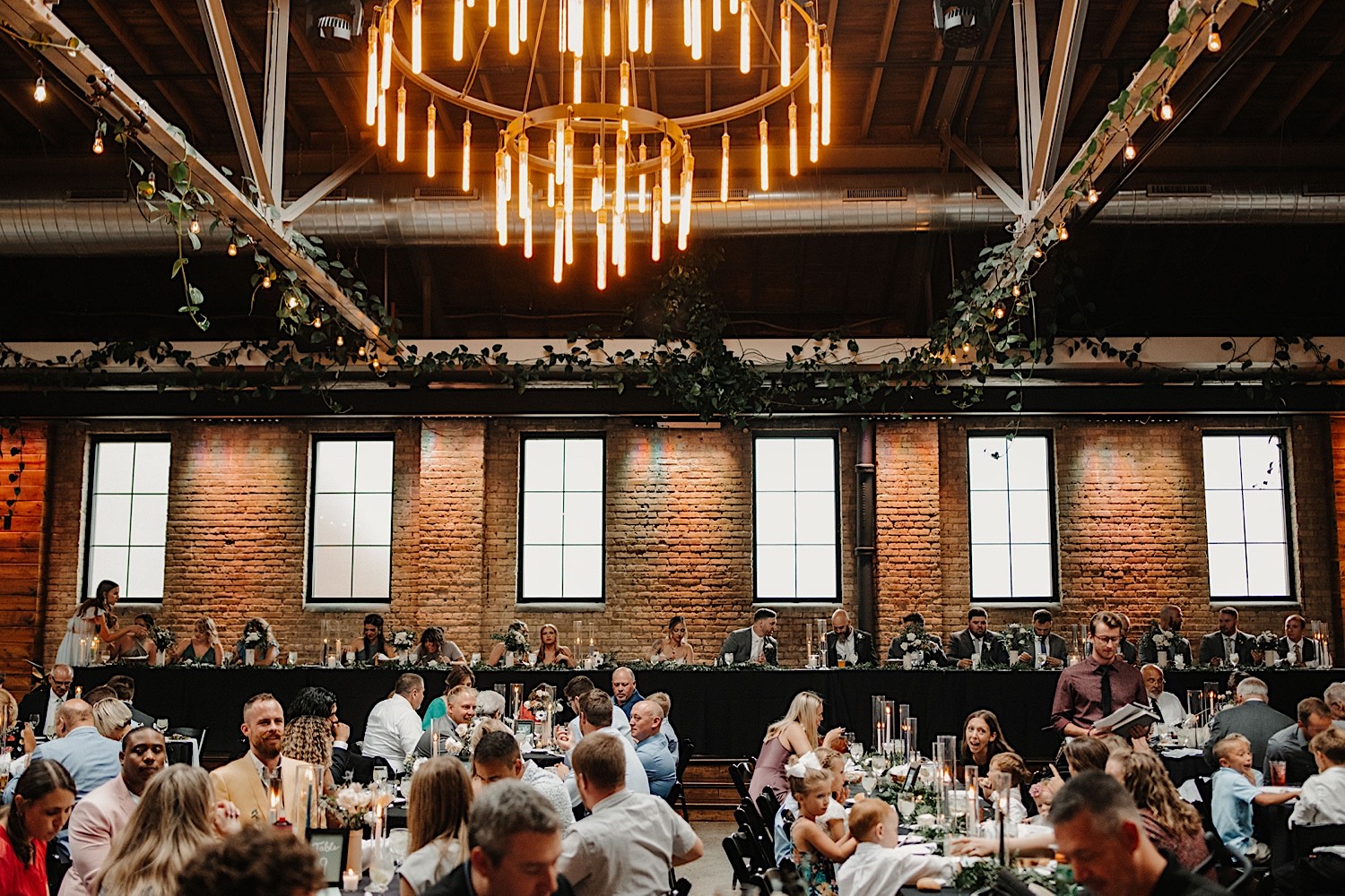 A wedding reception taking place at the wedding venue The Ivy House in Milwaukee, the reception is indoors and the head table is in front of a massive brick wall with ivy lining the rafters and a large chandelier overhead