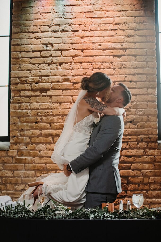 A bride is held and lifted in the air by the groom as the two kiss in front of a brick wall while at the head table of their reception