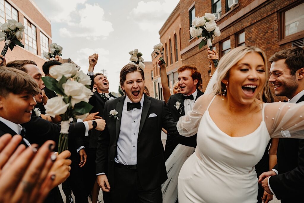 A bride runs and smiles towards the camera while holding the grooms hand who is also smiling and yelling behind her as their wedding parties surround them and cheer for them on either side on a street with brick buildings on it
