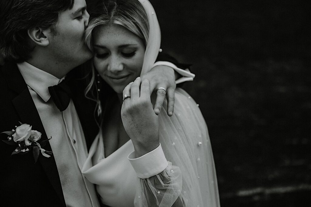 Black and white photo of a bride smiling while holding the hand of the groom who has his arm wrapped around her and is kissing her on the head