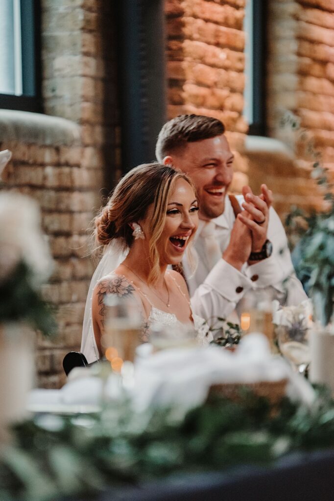 A bride and groom laugh and smile while sitting at their head table at something off camera