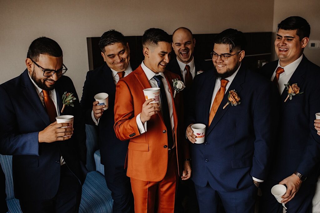 A groom smiles with his group of 6 groomsmen around him, each of them has a paper Dunkin cup in hand with liquor in it
