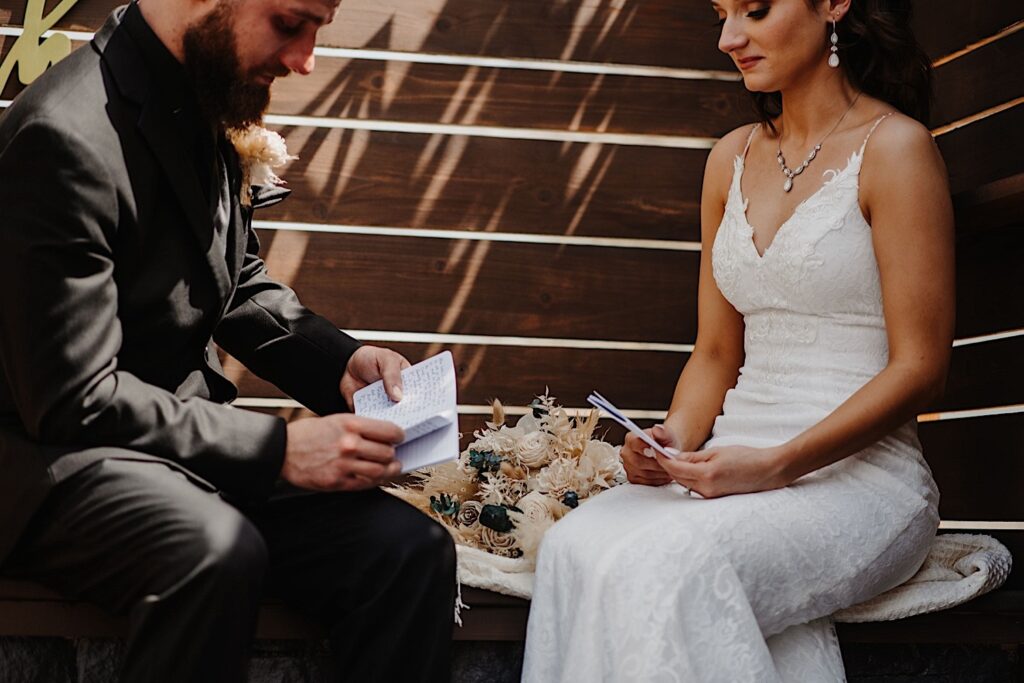 A bride and groom sit on a wooden bench with the bouquet between them, each with their wedding vows in their hands as they read them to one another