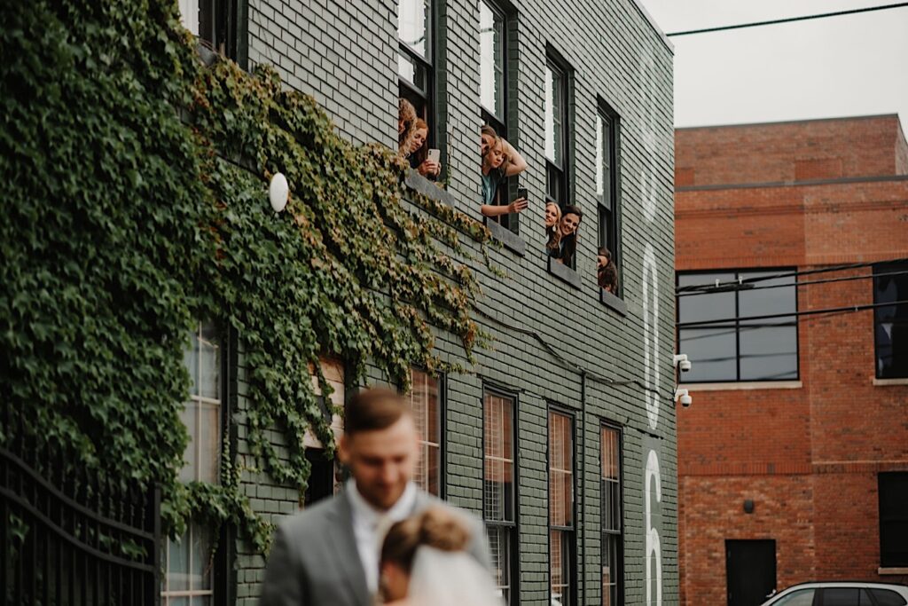 Bridesmaids in the second floor of the wedding venue The Ivy House all stick their heads out of the window to see the bride and groom who are outside on the ground level having their first look, the bride and groom are out of focus in the left foreground of the photo