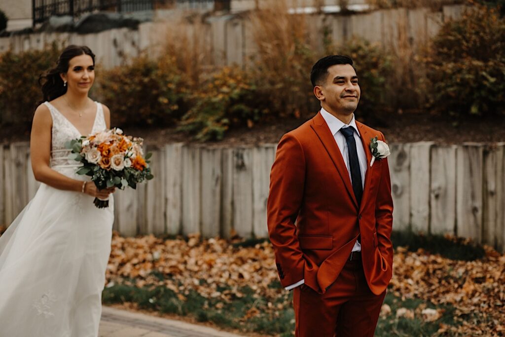 During their Fall Wedding at Mistwood Golf Club a groom smiles with his hands in his pockets with the bride standing behind him before their first look at outside their venue