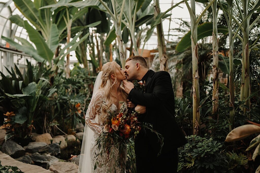 A bride and groom kiss one another inside of a greenhouse in front of large plants, the bride is holding a floral bouquet and the man has his hand lifting up the woman's chin