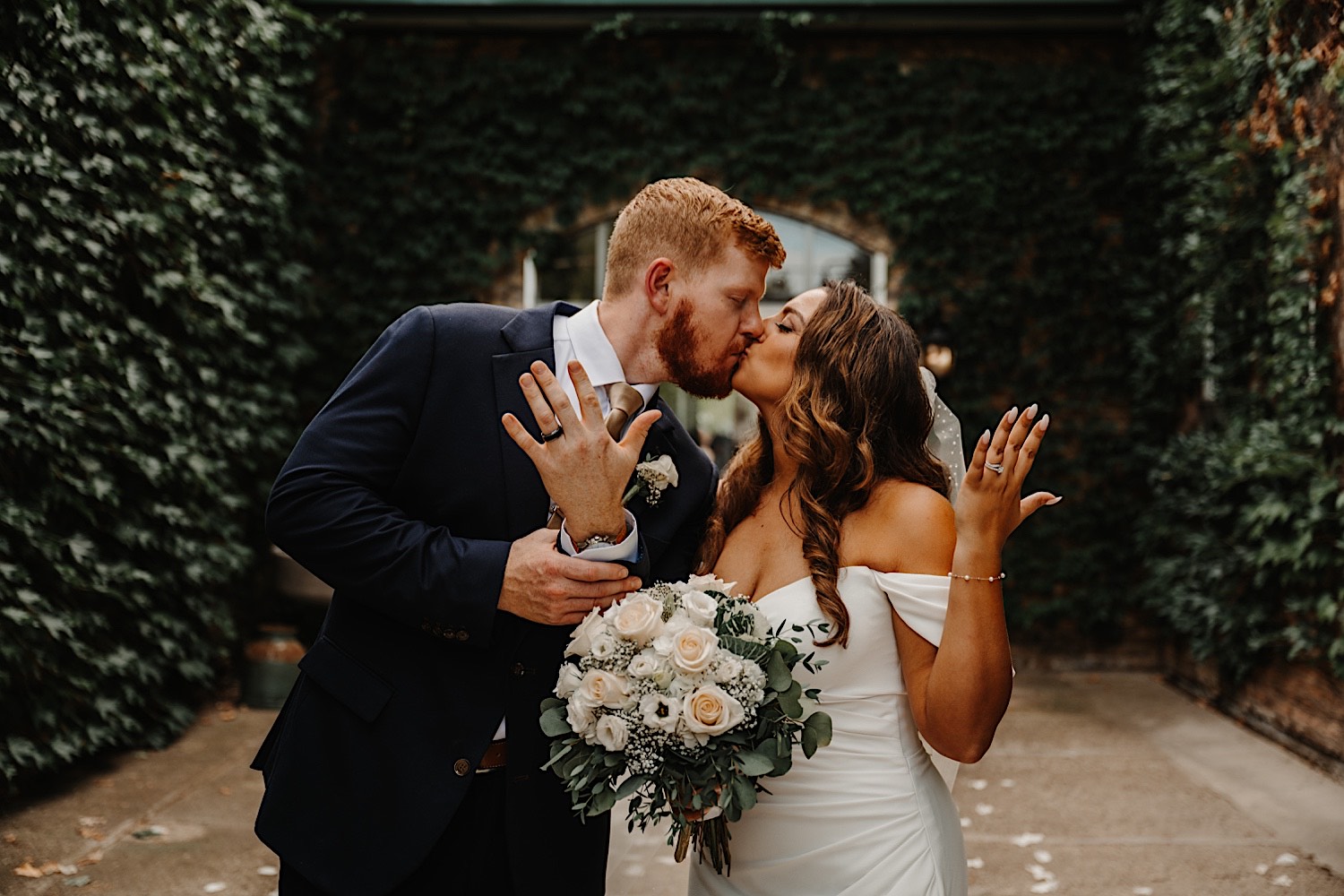 A bride and groom stand next to one another and kiss each other while holding out their hands with their wedding rings towards the camera while outside surrounded by walls covered in ivy