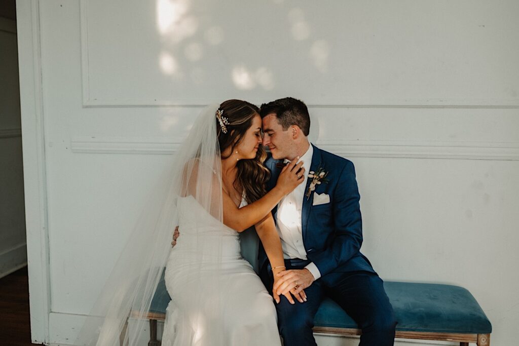 A bride and groom sit on a blue bench in front of a white wall and smile at one another while touching noses and their foreheads together with their eyes closed, the woman's hand rests on the man's chest and her other hand is being held by his on his lap