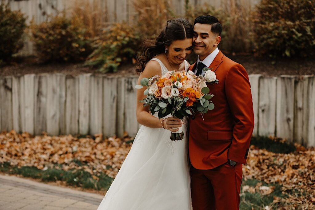 A bride and groom stand side by side and embrace while smiling outside their wedding venue Mistwood Golf Club with fall leaves behind them