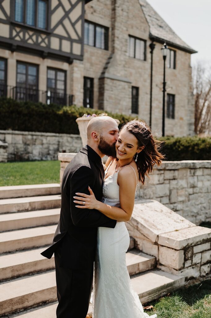 A bride smiles as the groom kisses her on the cheek in front of a stone staircase and building