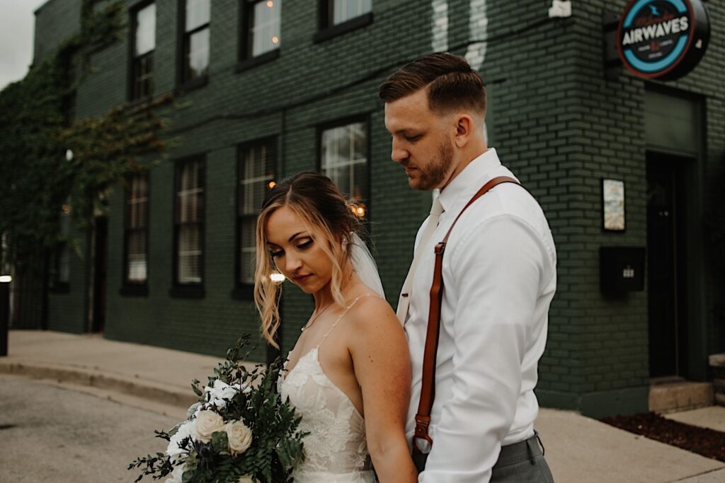 Outside of their wedding venue The Ivy House in Milwaukee, a bride stands in front of a groom who is holding her hands and looking down towards her as she looks down towards the ground