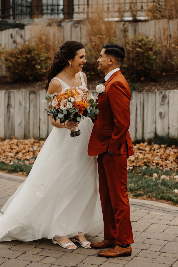 A bride and groom stand facing one another and smile while standing on a sidewalk with fall leaves next to it