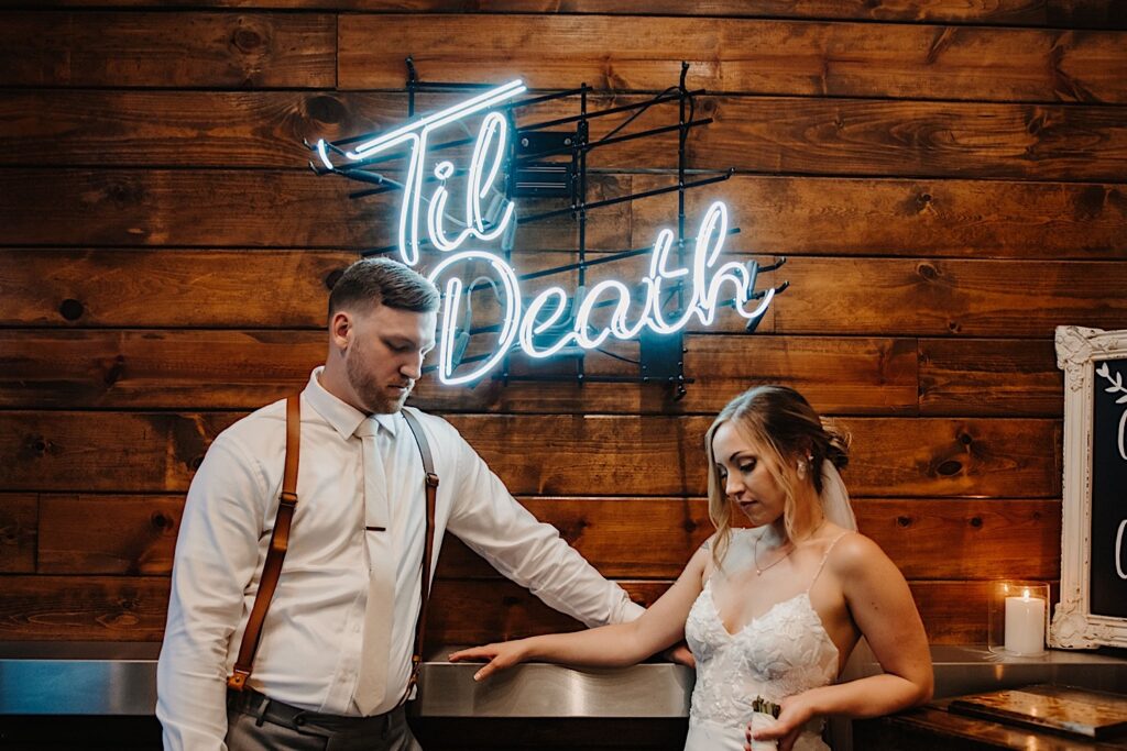 A bride and groom pose at a metal bar with a neon sign in between them that reads "Til Death" mounted on a wooden wall