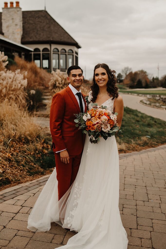 A bride and groom stand side by side and smile at the camera while outside a building with fall colors around them