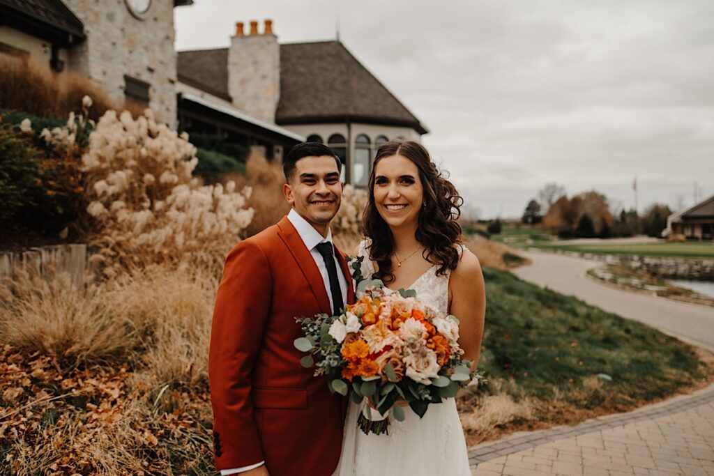 A bride and groom stand side by side and smile at the camera while outside their wedding venue Mistwood Golf Club with fall leaves and fall colored plants behind them
