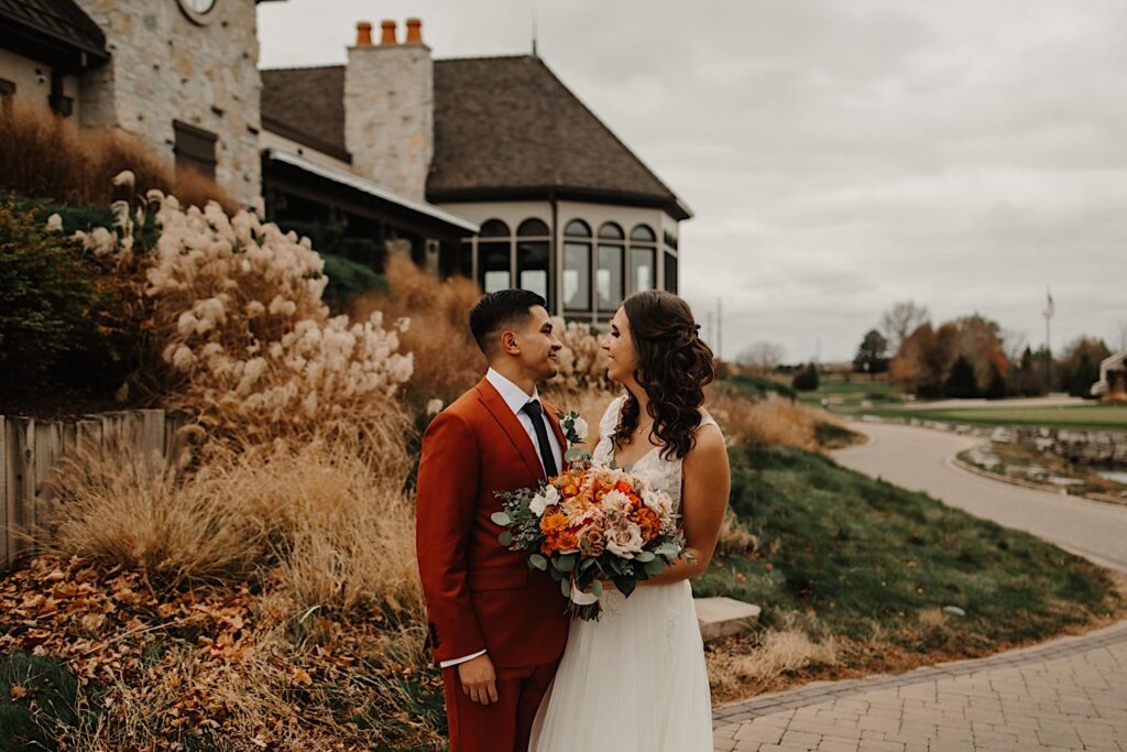A bride and groom stand next to one another and smile at each other while outside their wedding venue Mistwood Golf Club with fall leaves and fall colored plants behind them