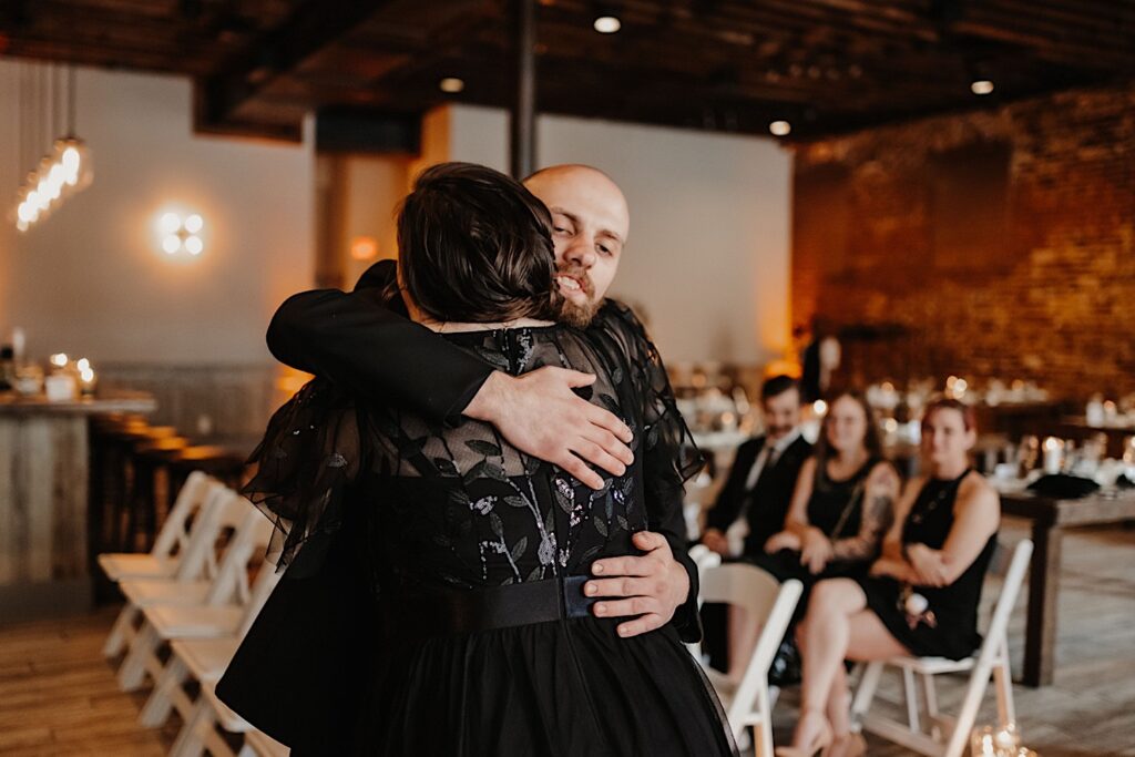 A groom smiles while hugging a guest of his wedding inside of Reality on Monroe's ceremony space as guests behind them watch