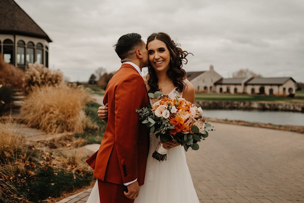 A bride smiles while the groom stands beside her and kisses her on the cheek while they stand outside their wedding venue Mistwood Golf Club with fall leaves and fall colored plants around them