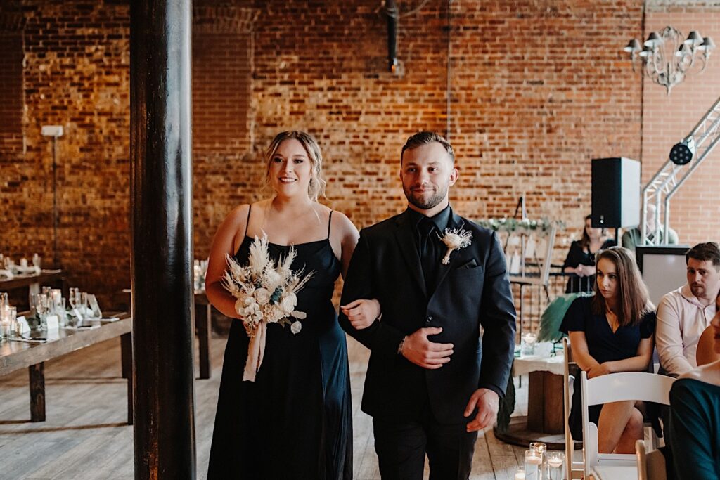 A bridesmaid and groomsmen smile while walking down the aisle together for a wedding ceremony at Reality on Monroe as guests on their right watch