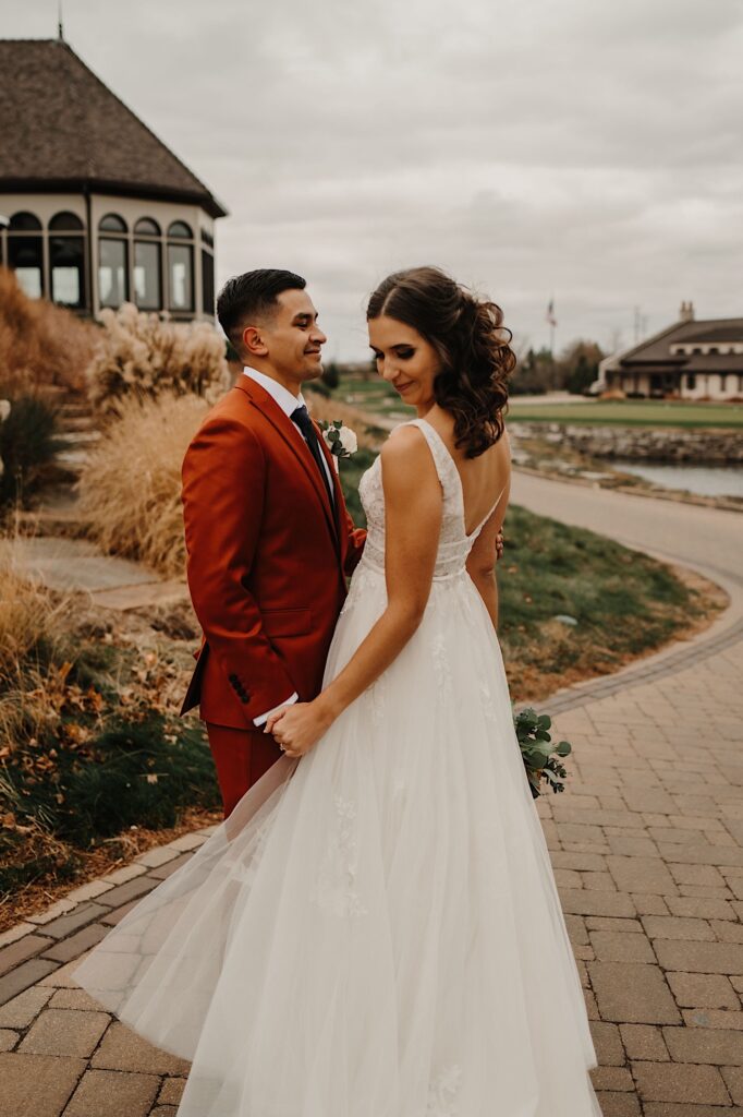 A bride and groom face one another and hold hands as the bride looks down at her hand, the two are on a sidewalk next to a building in the fall
