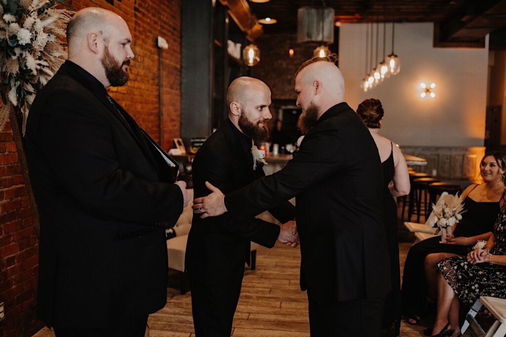 A groom smiles while shaking the hand of one of his groomsmen next to the officiant of his wedding ceremony taking place at Reality on Monroe