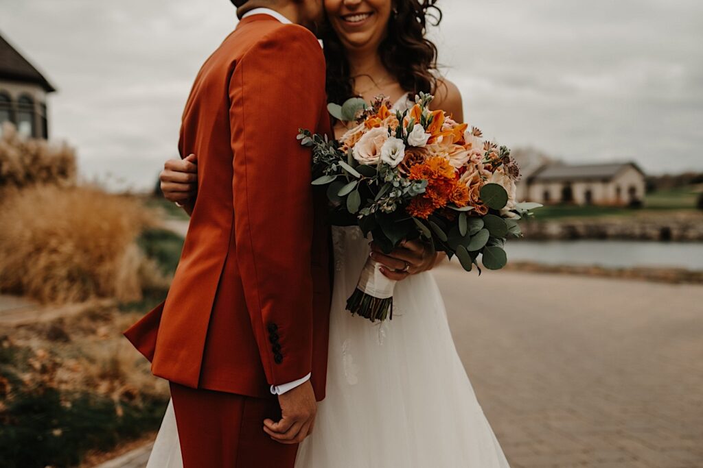A photo of a bride smiling while the groom stands beside her and kisses her on the cheek while they stand on a sidewalk outside their wedding venue Mistwood Golf Club in the fall