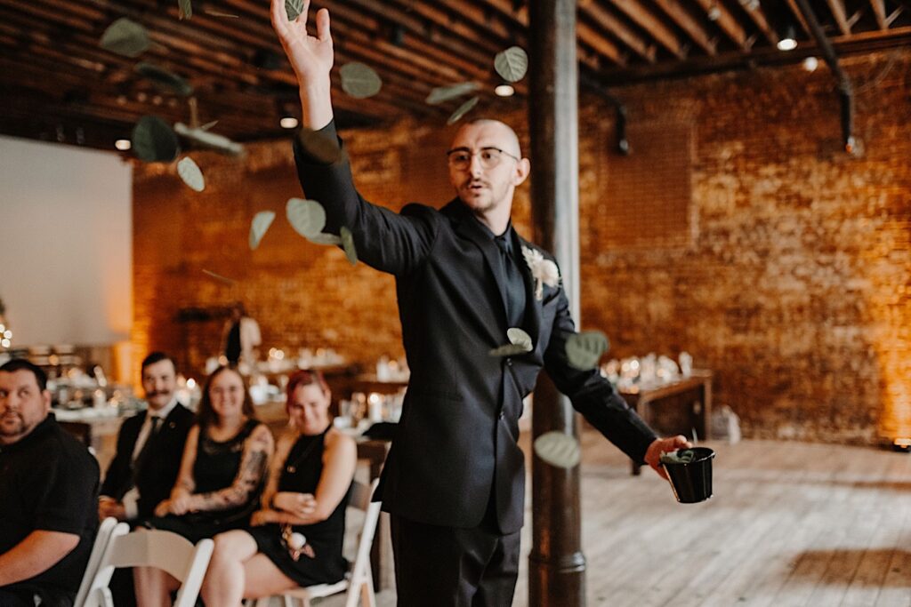 A groomsmen acting as the flower boy throws leaves in the air while walking down the aisle as guests watch and laugh during a wedding ceremony at Reality on Monroe