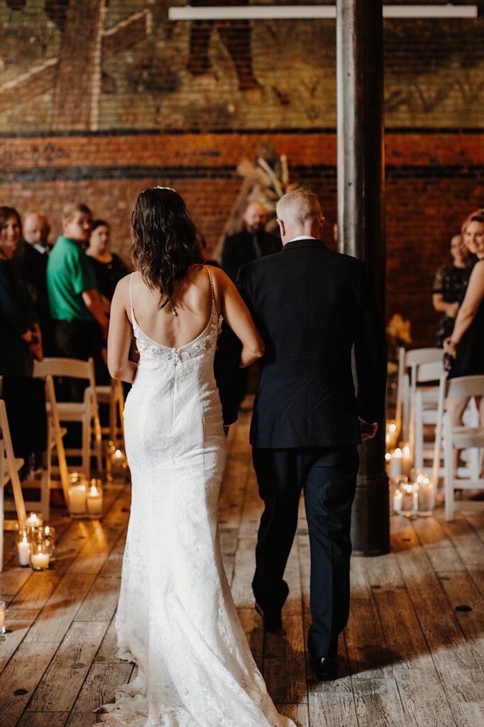 A bride and her father walk away from the camera down the aisle of a wedding ceremony as guests on either side watch and smile