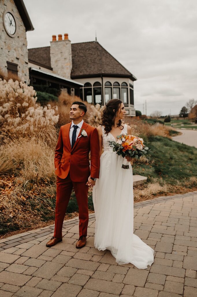 A bride and groom stand side by side and look in opposite directions while on a sidewalk outside a building in the fall