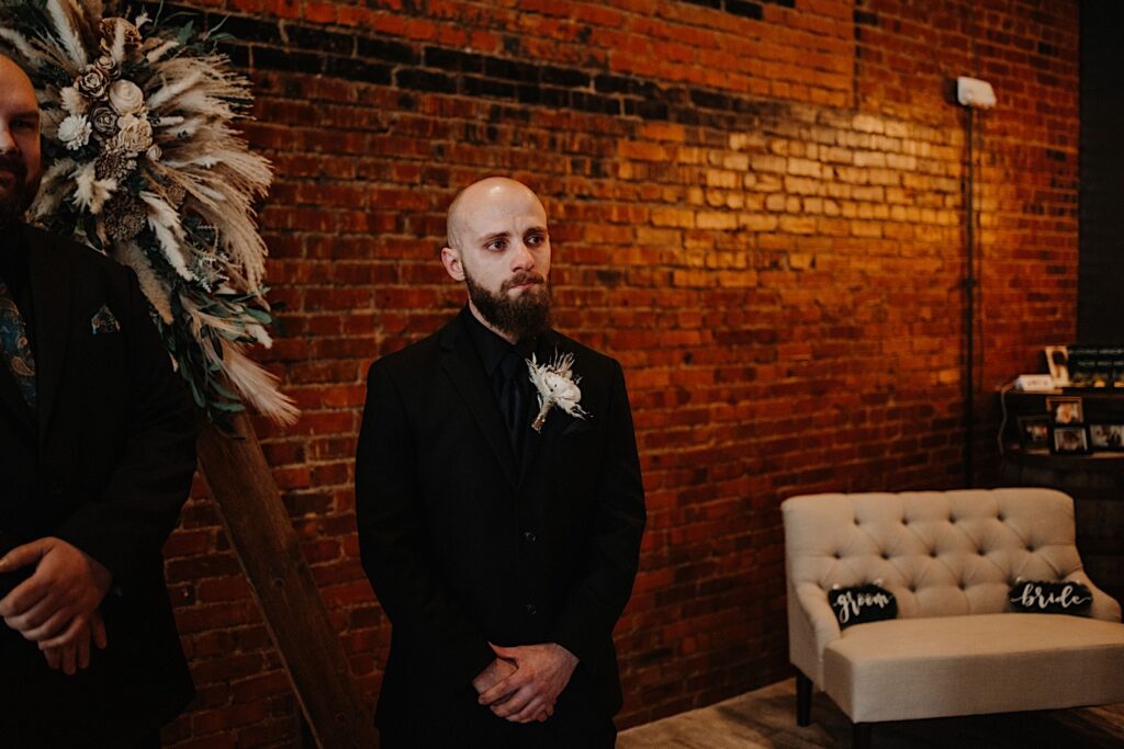 A groom gets emotional standing in front of a brick wall inside Reality on Monroe as he sees the bride walking down the aisle towards him during their wedding ceremony