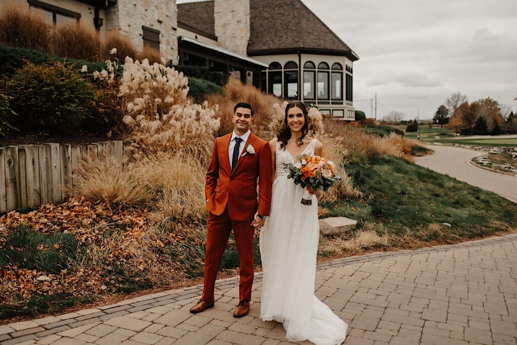A bride and groom smile at the camera while holding hands and standing next to one another while on a sidewalk outside their wedding venue Mistwood Golf Club in the fall