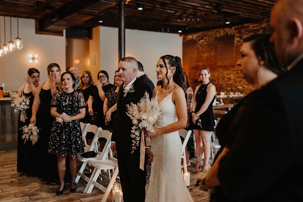 A bride smiles while holding her bouquet and being walked down the aisle by her father as guests watch and tear up during a wedding ceremony at Reality on Monroe