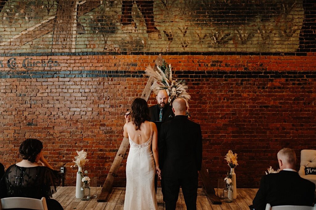 A bride and groom stand side by side holding hands facing away from the camera towards their officiant speaking while standing in front of a brick wall at Reality on Monroe's indoor wedding ceremony space