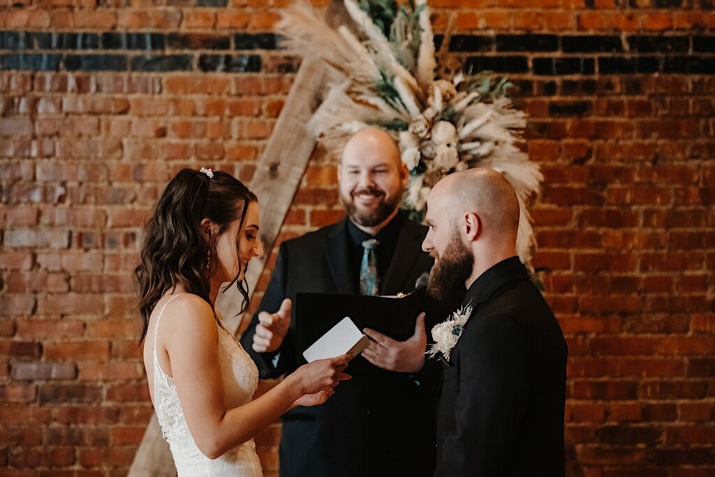 A bride smiles while reading her vows to the groom who is smiling back at her, in the background their officiant smiles while standing in front of a brick wall in Reality on Monroe's indoor wedding ceremony space
