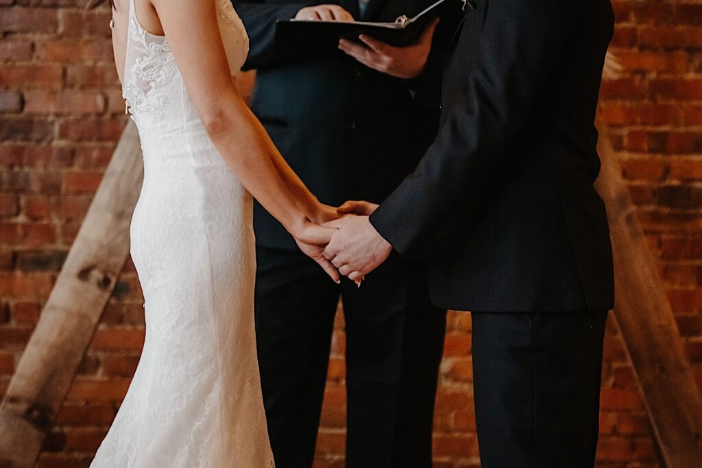 Shoulder down photo of a bride and groom holding hands in front of their officiant and a brick wall
