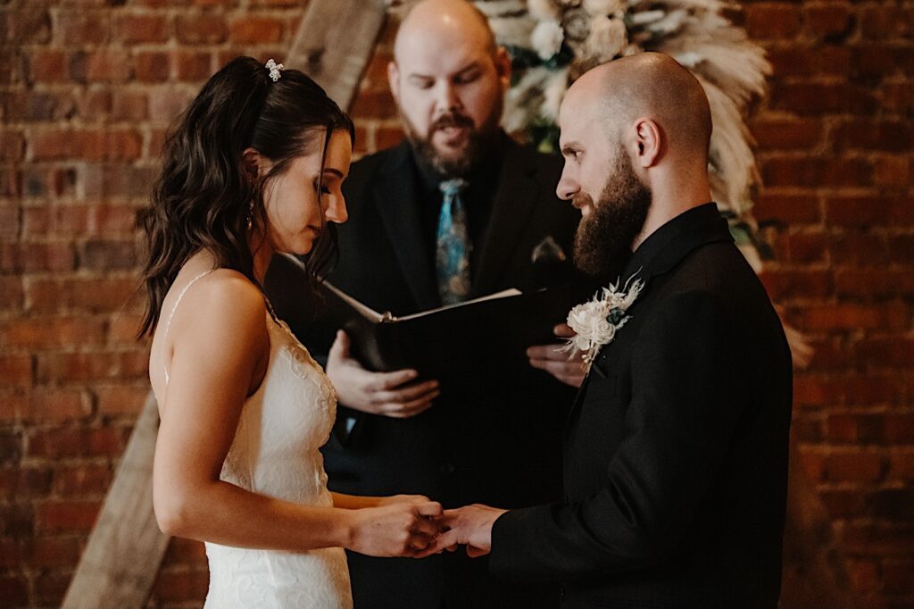 An officiant speaks while the bride puts a ring on the grooms hand as he looks at her during their indoor wedding ceremony at Reality on Monroe