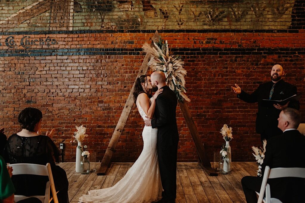 A bride and groom kiss in front of a brick wall and their wooden archway, to the right their officiant speaks to the crowd as they clap and watch, the wedding ceremony is taking place inside Reality on Monroe in Bloomington