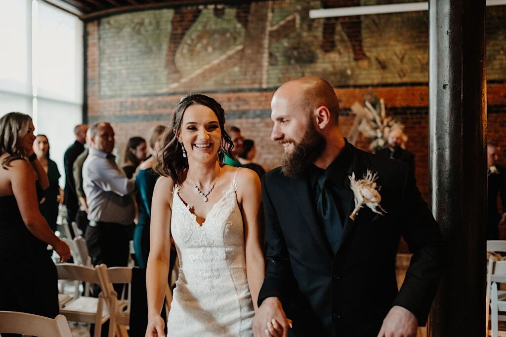 A bride smiles at the camera while the groom smiles at her as they walk hand in hand down the aisle as guests clap for them after their indoor wedding ceremony at Reality on Monroe