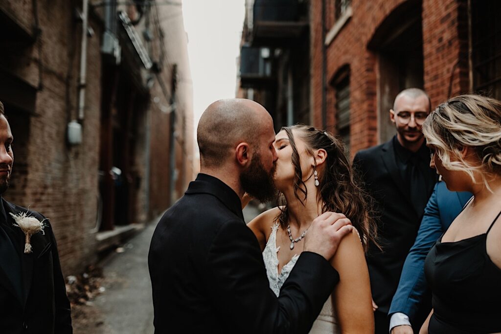A bride and groom kiss one another in an alley with their wedding party around them watching 