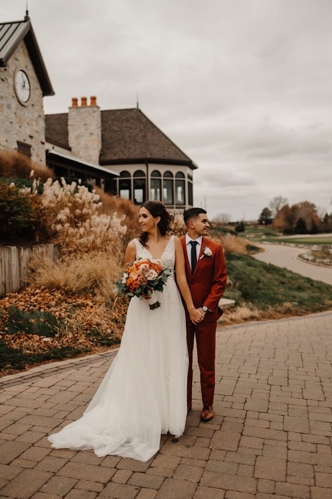 A bride stands in front of a groom and holds his hand while the two look in opposite directions while on a path next to a building in the fall
