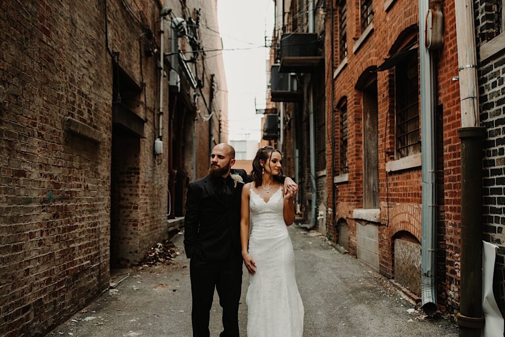A bride and groom stand in an alley outside of their wedding venue Reality on Monroe and look in opposite directions while the groom wraps his arm around the bride's shoulder