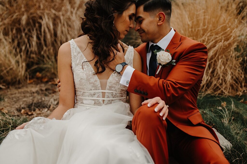 A bride and groom sit in the grass outside their wedding venue Mistwood Golf Club and are about to kiss one another with tall grass behind them turned brown from the fall weather