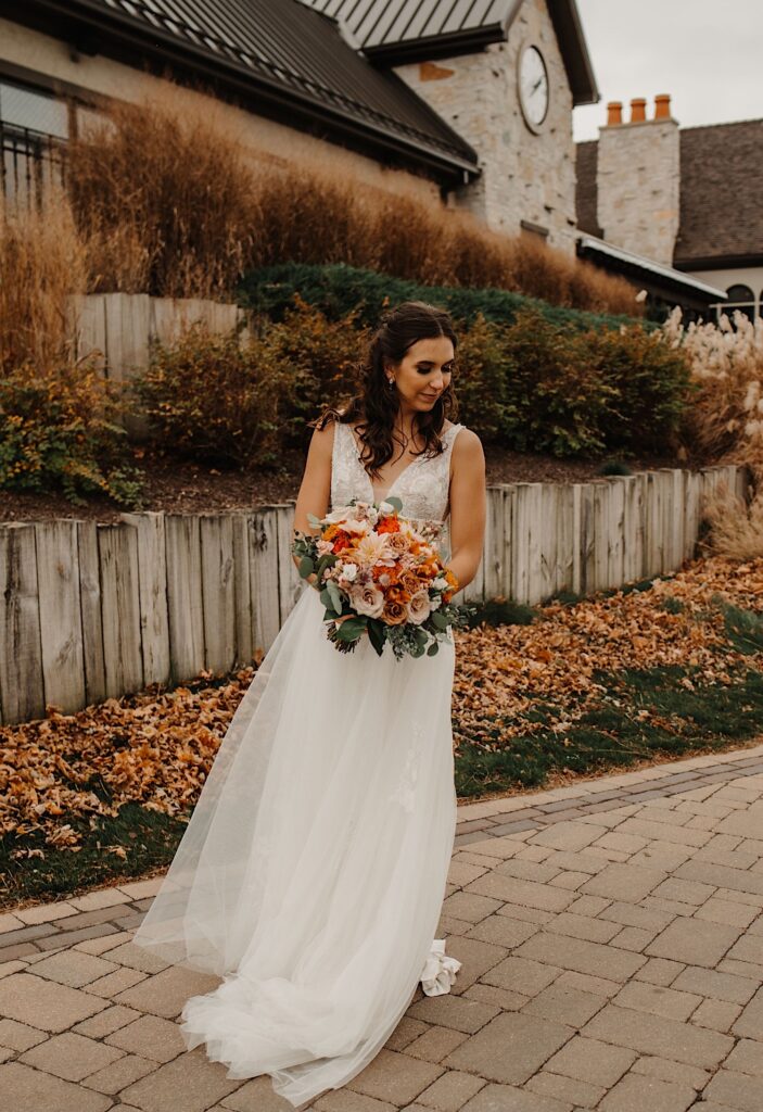 A bride stands on a path in front of a building with leaves on the ground behind her and smiles at the ground to the right while holding her bouquet