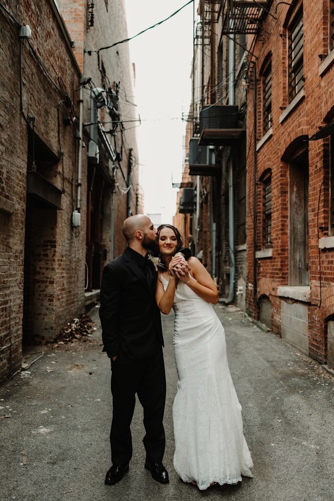 A bride smiles at the camera while the groom kisses her on the side of the head and wraps his arm around her shoulder as the two stand in an alley