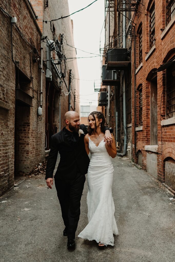 A bride and groom walk together down an alley and smile at one another while the groom wraps his arm around the bride's shoulder